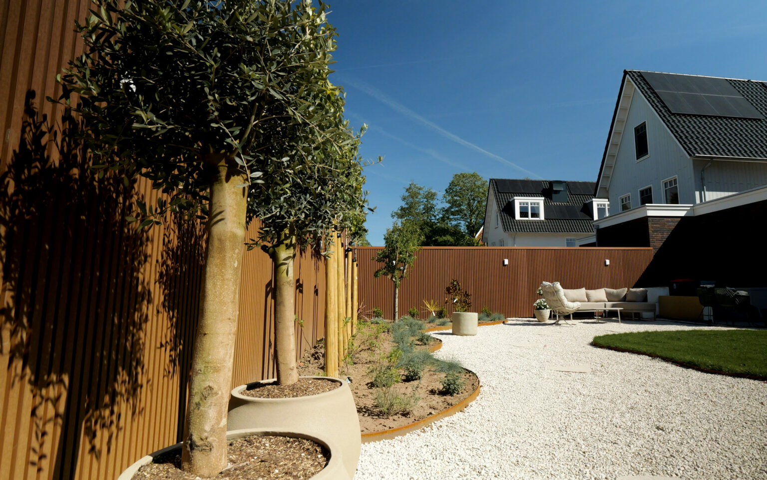 Moderne tuin met bruine houtcomposiet schutting, siergrind, cortenstalen borderranden en jonge mediterane beplanting.
